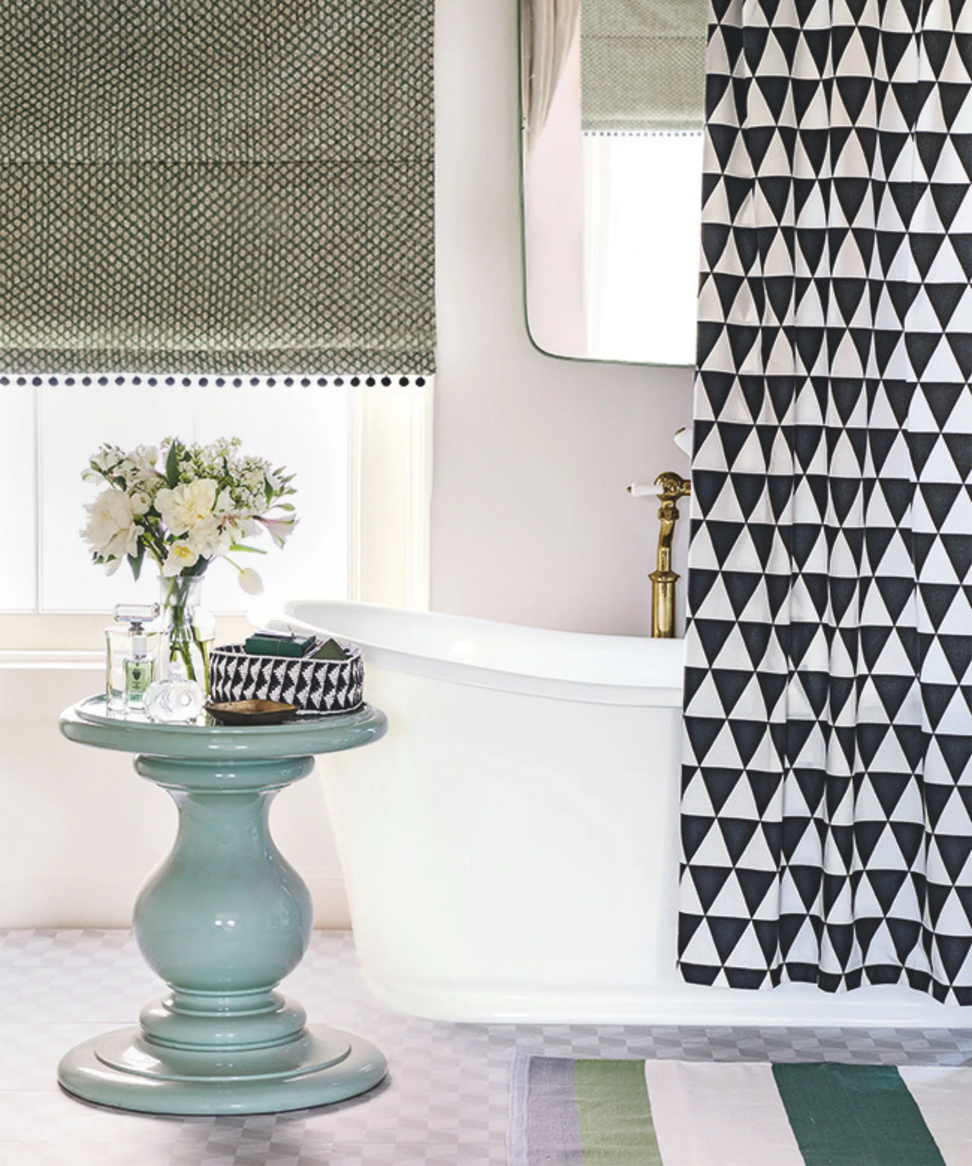 Una bañera blanca en un baño luminoso con una cortina de ducha en blanco y negro y un espejo con grifo dorado encima y una mesa circular azul con un jarrón transparente de flores blancas encima. Detrás hay un gran ventanal con persiana verde y en el suelo hay una alfombra de rayas verdes y blancas.' loading='lazy' title=