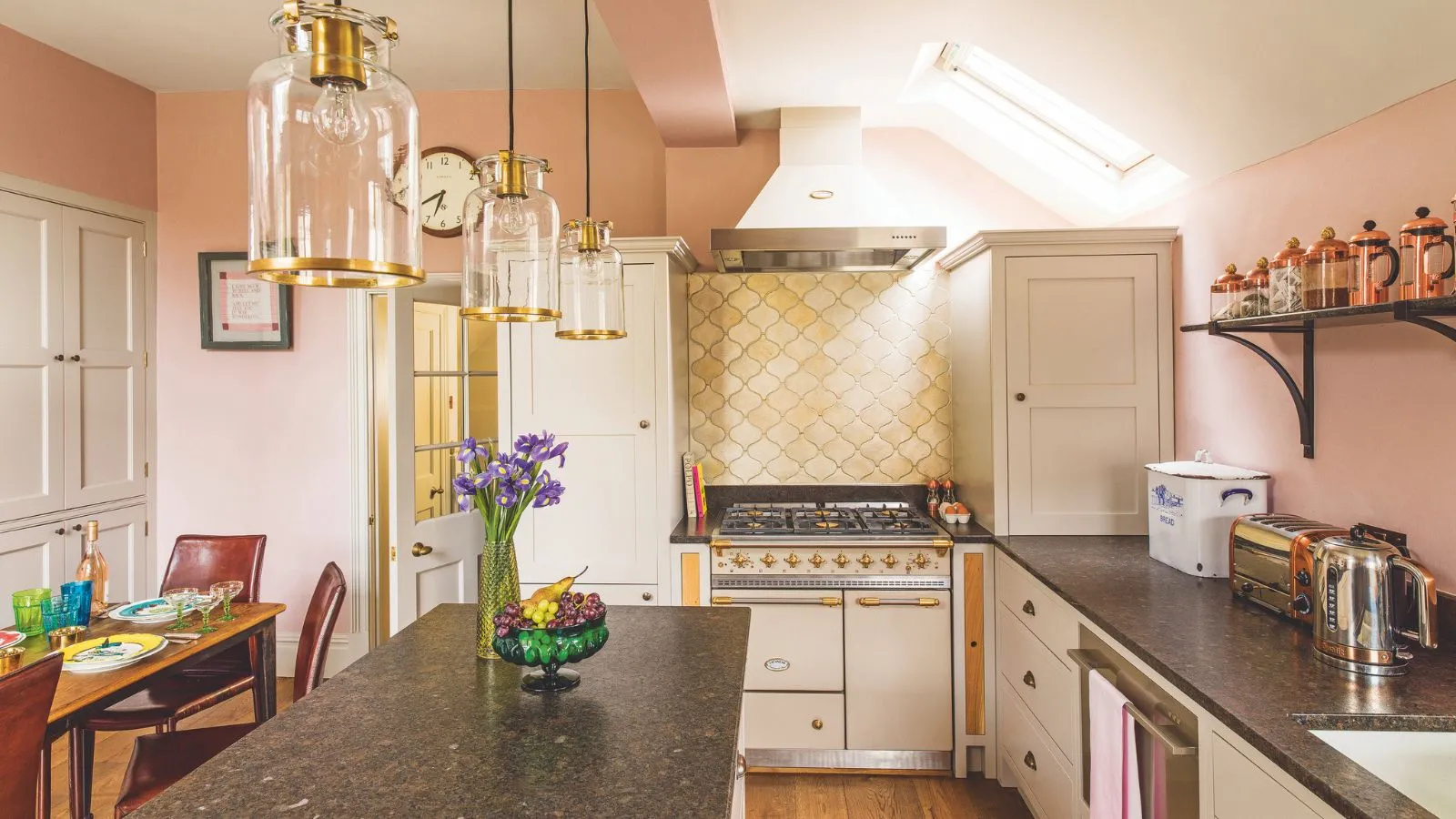 Cuisine rose kitsch avec dosseret jaune en forme d'étagère derrière une cuisinière crème et or. Il s'agit d'une cuisine-salle à manger avec un bar pour le petit-déjeuner et des suspensions aux accents dorés.' loading='lazy' title=