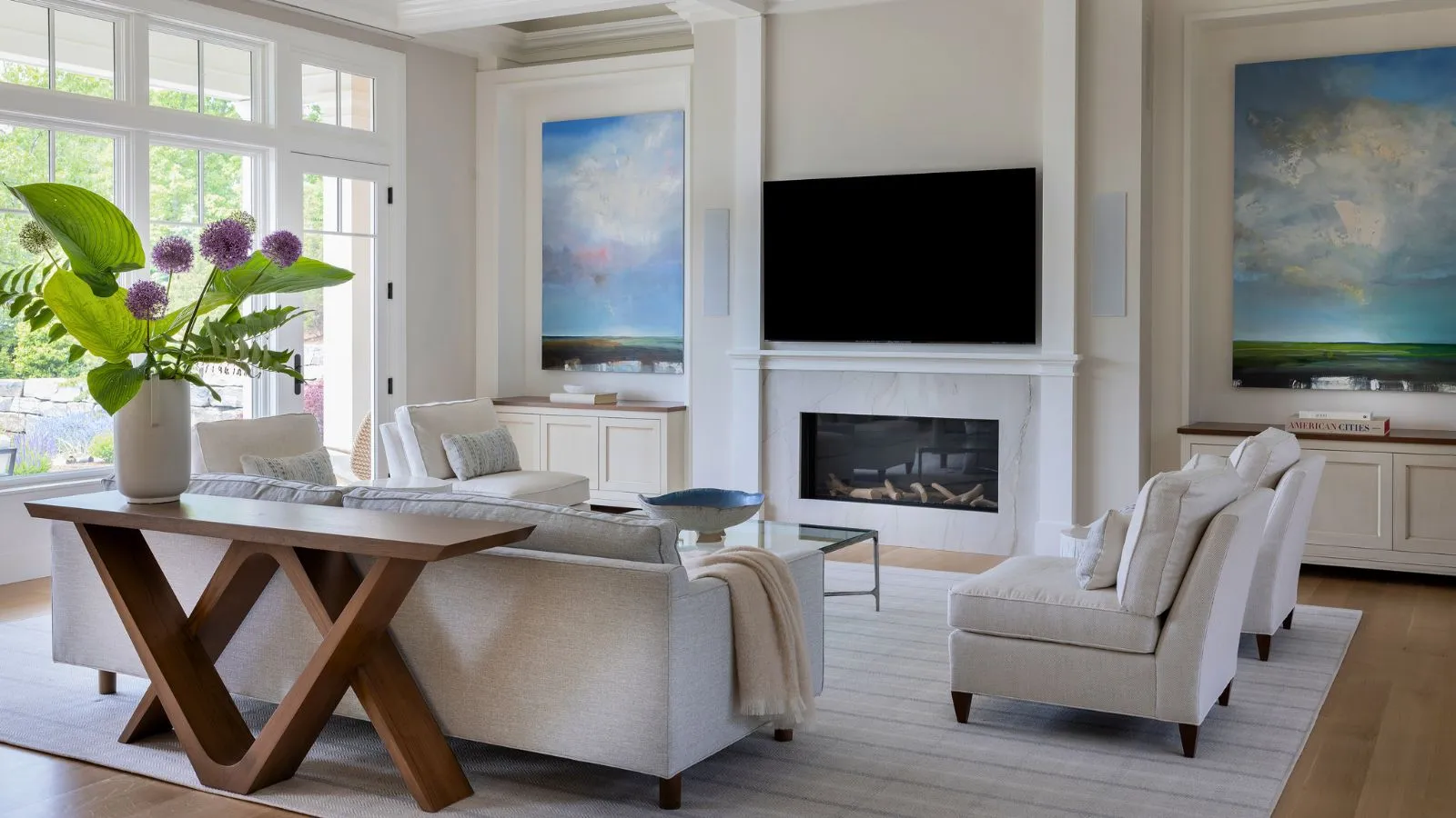 Hvid stue med højt til loftet pejs med monteret TV over hårde gulve og tæppe med sofaer indstillet til at centrere TV'et. Store vinduer giver masser af naturligt lys og en trækonsol i forgrunden viser enorme lilla blomster og store grønne blade' loading='lazy' title=