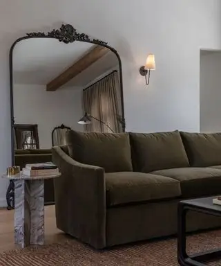 Veľké starožitné detailné podlahové zrkadlo opreté o stenu za olivovo zeleným gaučom s malým príručným stolíkom s mramorovou povrchovou úpravou a nástenným svietnikom