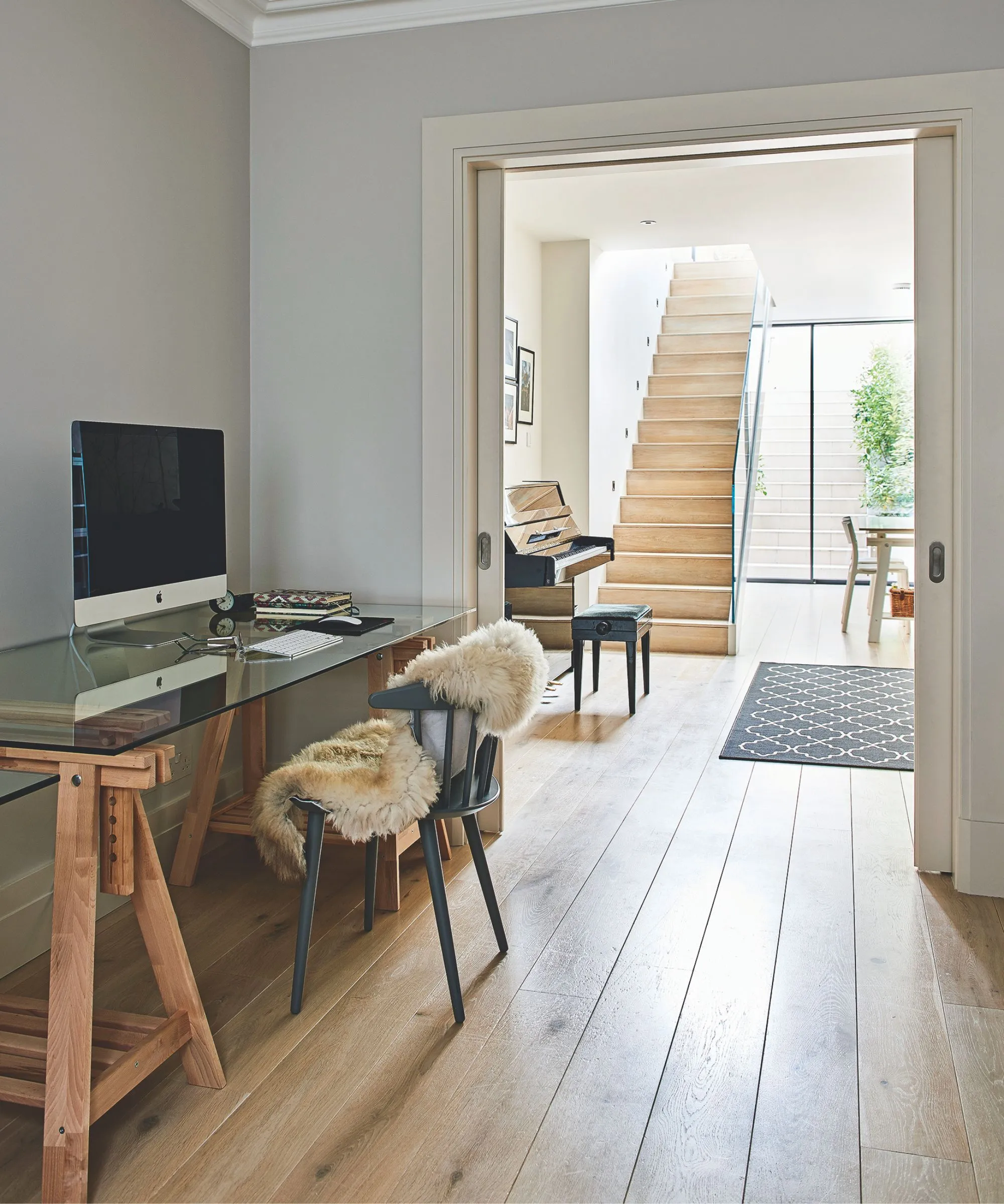 En lys og luftig kjeller med tregulv en tre- og glasspult med en trestol med et luftig kast og et piano i bakgrunnen tilstøtende rom med et svart mønstret teppe og tretrapp oppe. I bakgrunnen er glassdører ut til en hagetrapp med en løvgrønn plante til høyre.' loading='lazy' title=