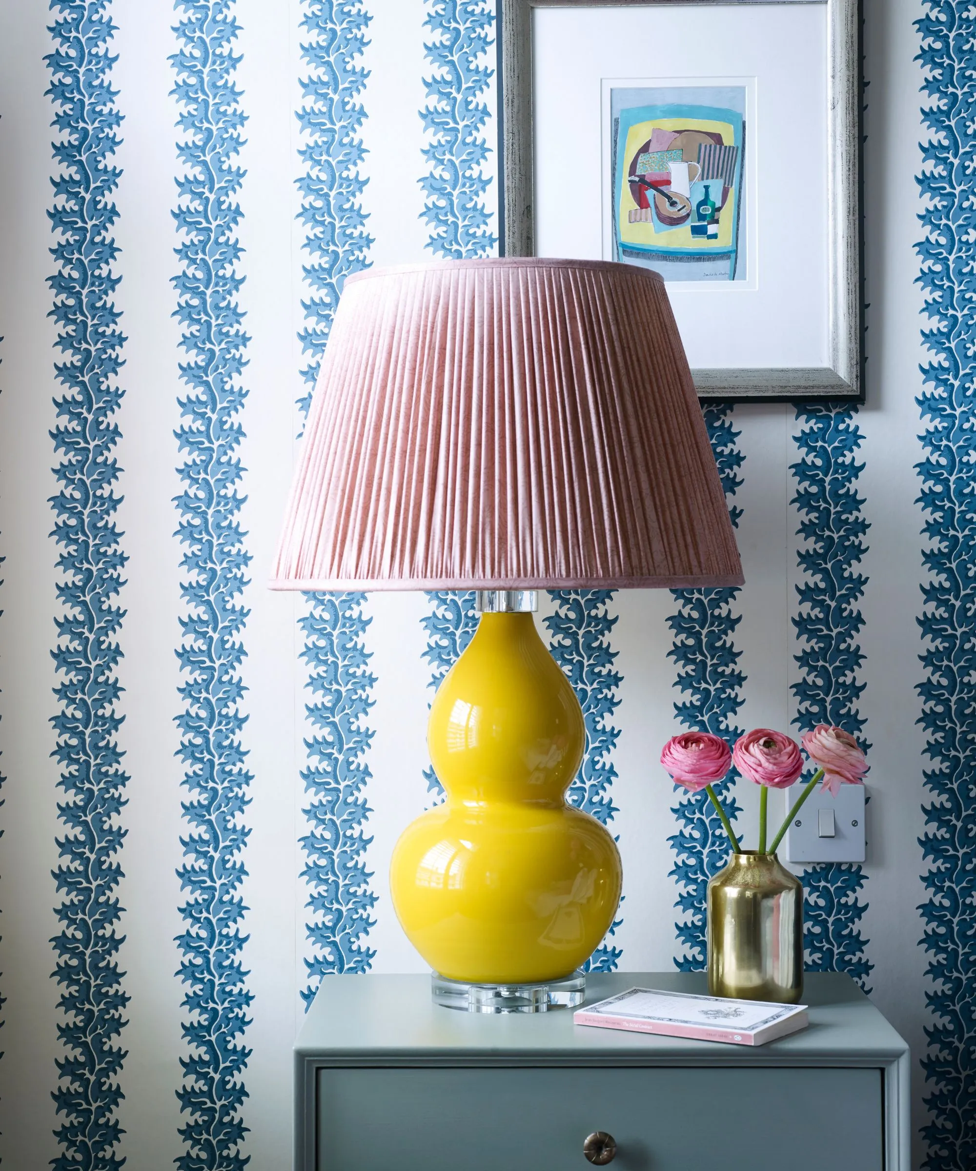 Et rom med blå og hvit statement tapet en gul keramisk lampe med rosa lampeskjerm innrammet kunstverk i sølvramme og blått nattbord med en gullvase fylt med rosa blomster og en bok.' loading='lazy' title=