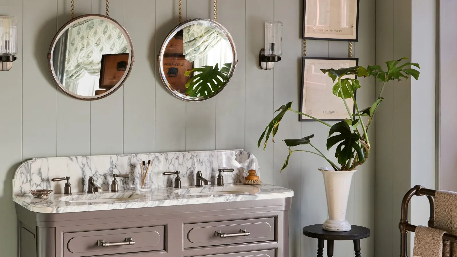 Sage Green Paneled Bathroom with Cream og Gray Veined Marble Double Vanity. Det er to runde speil som henger fra kjeder over hver vask og vintage vegglamper' loading='lazy' title=