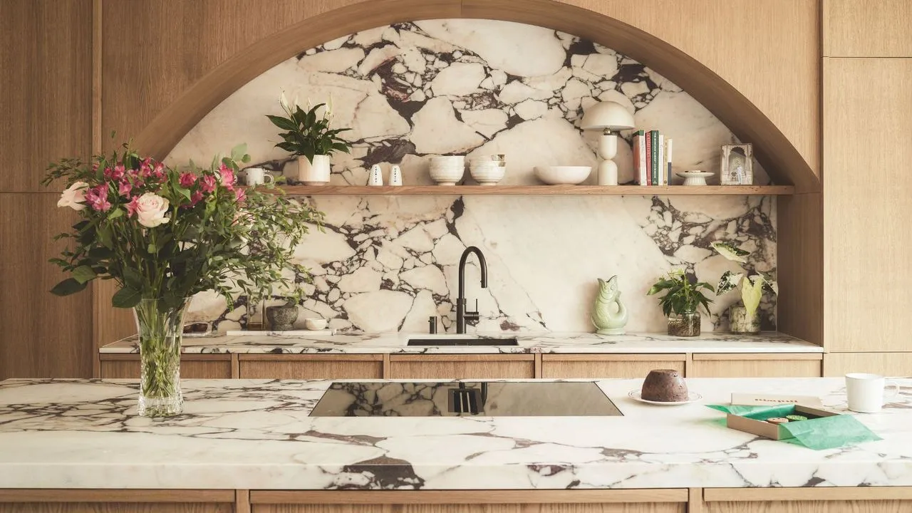 大きな大理石のバックスプラッシュを備えたキッチン シンクの上に、アーチ型のライトウッドの金具を備えたモダンなキッチン。向かい側のアイランドには、大理石のトップとガラスのコンロトップがマッチしています。左側にはバラの花瓶。' loading='lazy' title=