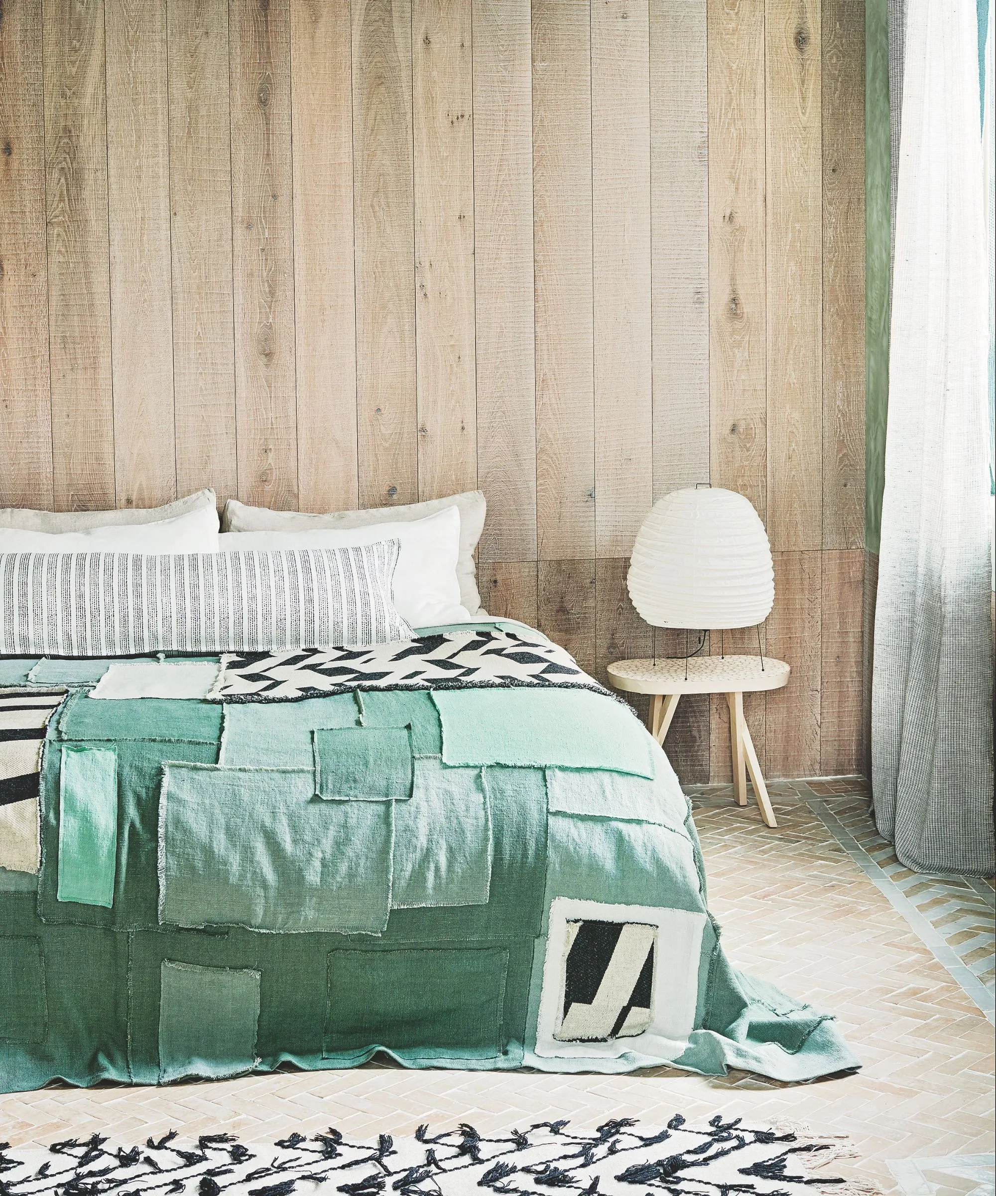 Una camera da letto con pannelli in legno chiaro con pavimento piastrellato e un letto matrimoniale con trapunta patchwork verde, cuscini bianchi, una sedia bianca e un tappeto bianco e nero.' loading='lazy' title=