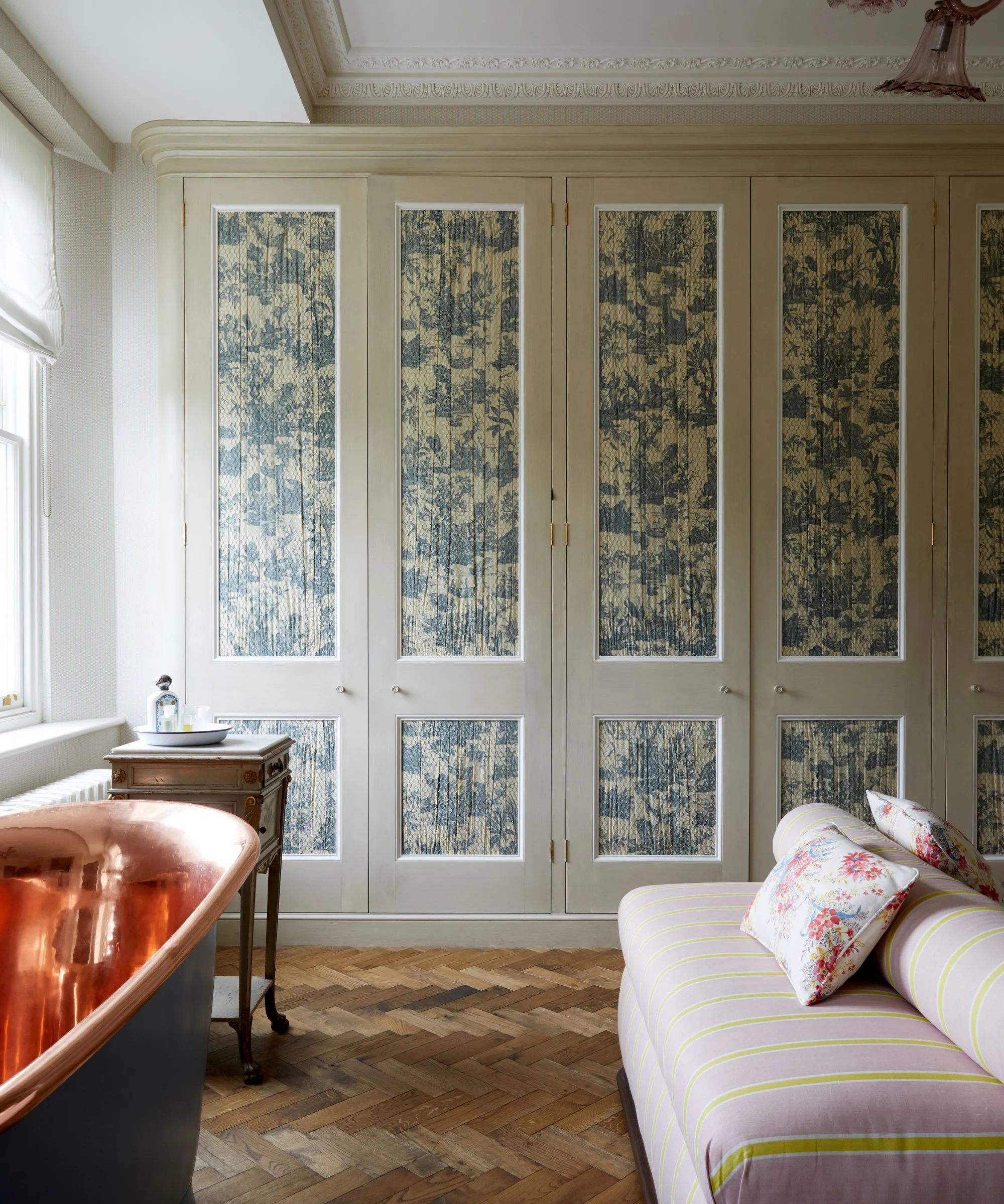 Un armadio dai motivi particolari con pavimento in parquet, un tavolo in legno con vasca da bagno in rame e nero e un divano a strisce rosa e gialle con cuscini floreali.' loading='lazy' title=