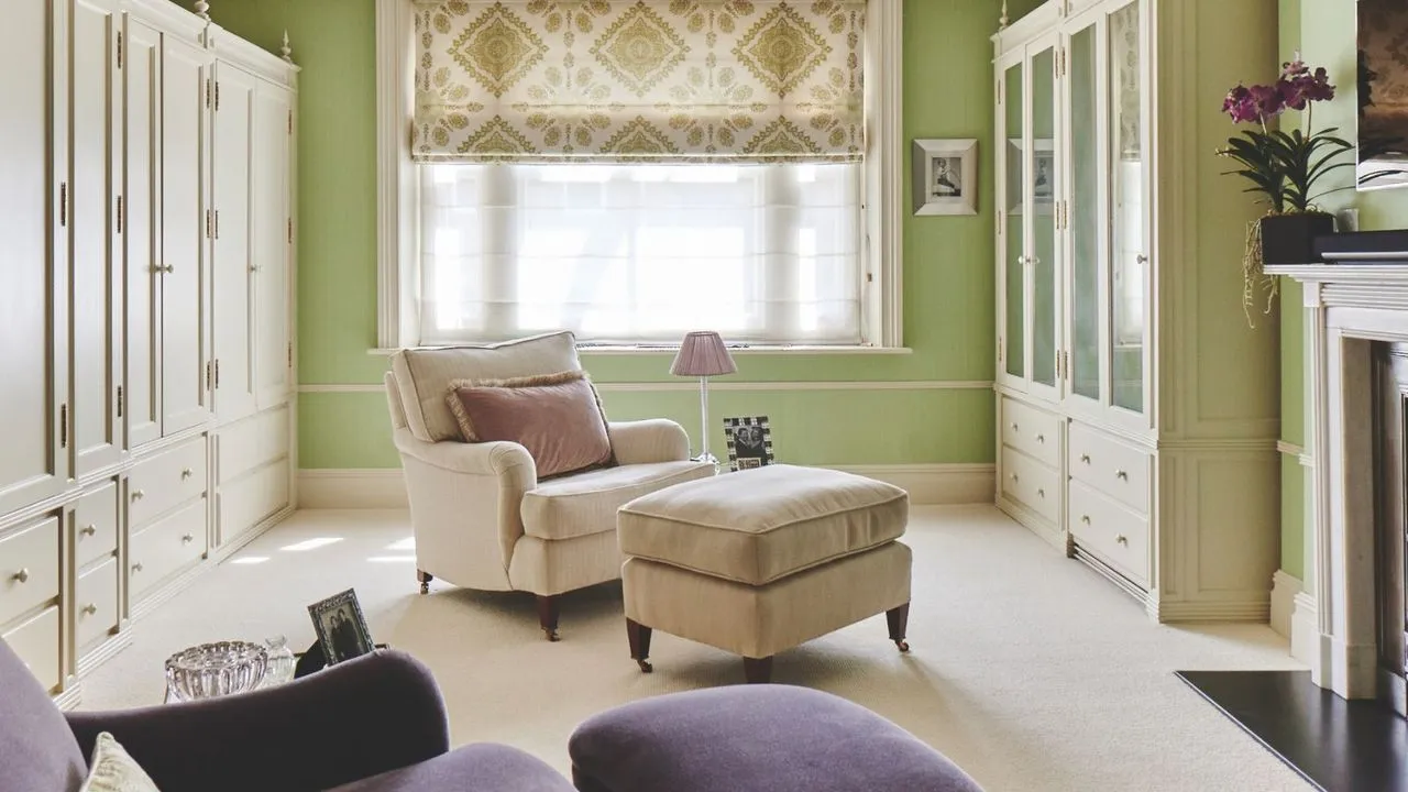 Uno spogliatoio verde con moquette bianca, armadi a muro bianchi, una grande finestra con una tenda trasparente e una tenda a motivi verdi e bianchi e un caminetto bianco sulla destra. Al centro della stanza c'è una sedia imbottita bianca con poggiapiedi abbinato, lampada e cuscino rosa e in primo piano c'è una sedia imbottita viola.' loading='lazy' title=