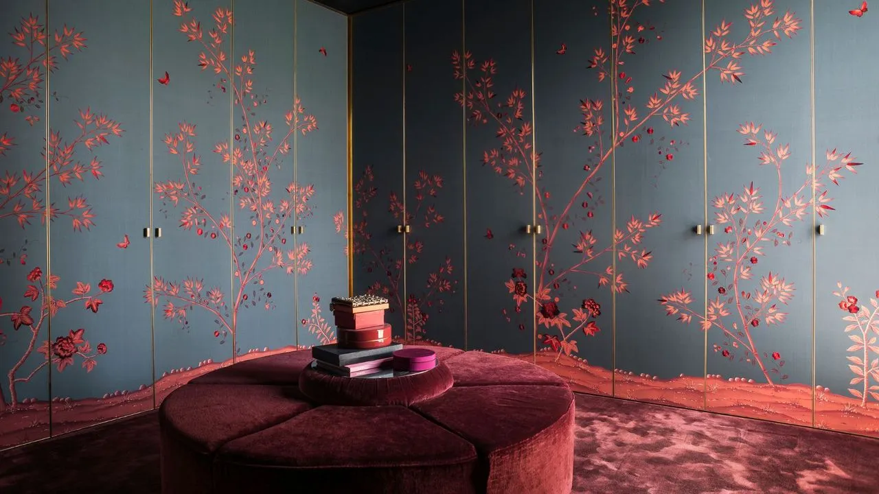 Cabina armadio di lusso scuro con armadi a muro dal pavimento al soffitto, ante tappezzate di blu scuro opaco con disegno floreale orientale rosso. Lussuosi tappeti bordeaux con velluto rotondo abbinato al grande sedile al centro' loading='lazy' title=