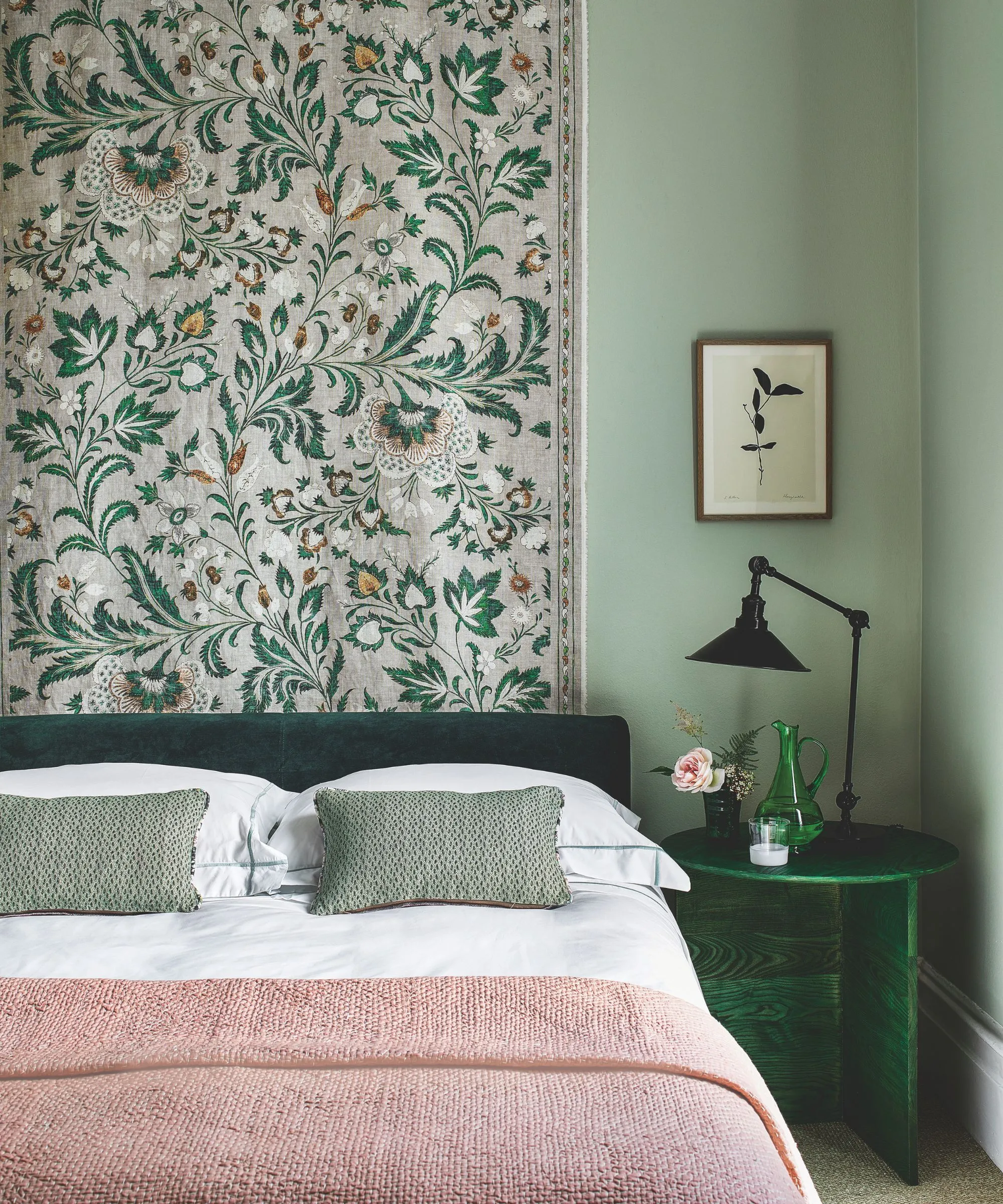 Ein grünes Schlafzimmer mit einem grünen Samtbett mit weißer Bettwäsche, einer rosa Decke und grünen Kissen, einem gemusterten Wandteppich hinter dem Bett, einem grünen Nachttisch mit einem grünen Glaskrug, rosa Blumen und einer schwarzen Lampe mit einem kleinen botanischen Kunstwerk darüber. Die Wände sind in sattem Salbeigrün gehalten.' loading='lazy' title=