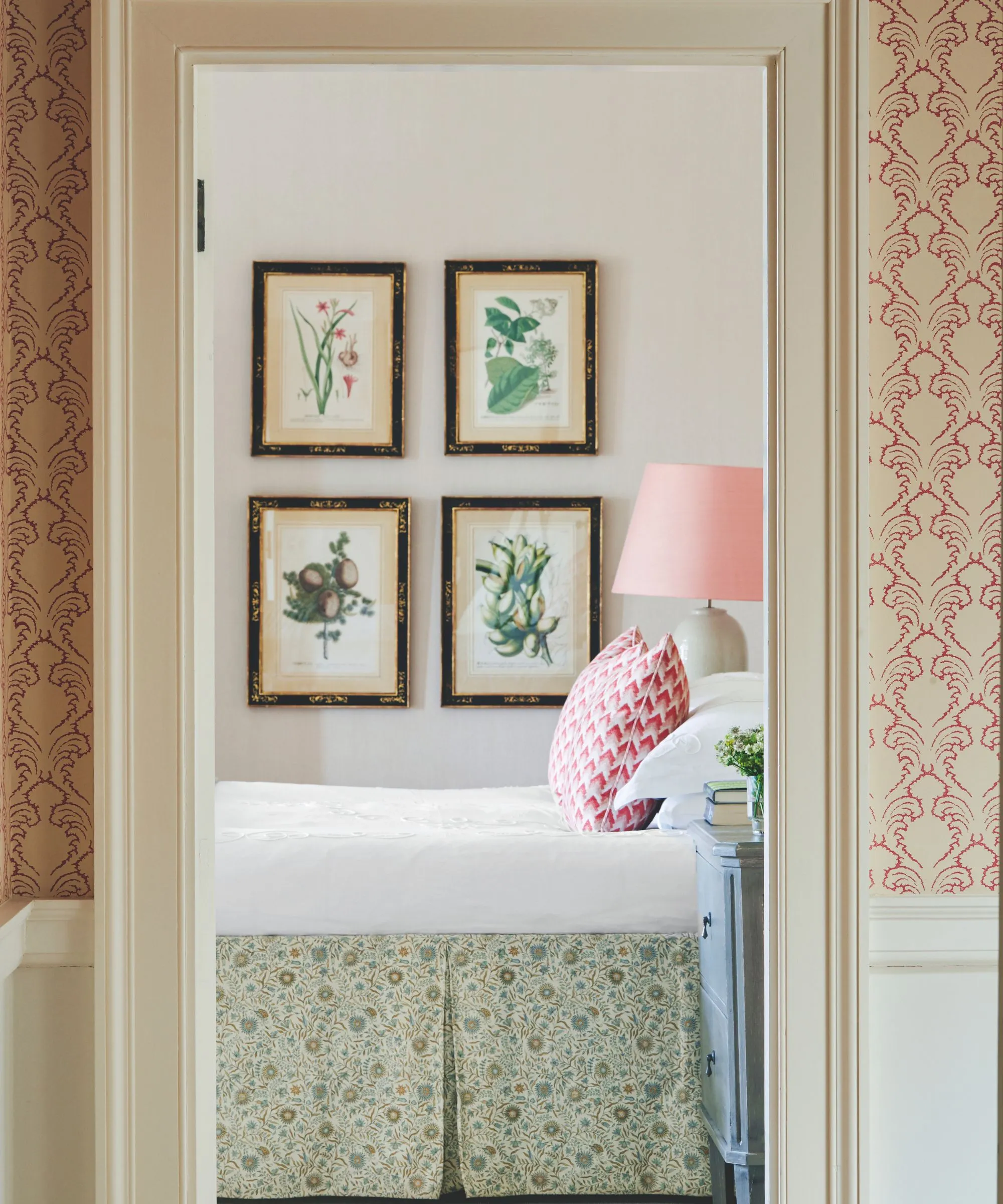 Eine weiße Tür mit rosa-weißer Tapete auf beiden Seiten und weißen Fußleisten gibt den Blick in ein Schlafzimmer mit einem Bett mit weißer Bettwäsche, einem Bettrock mit grünem Blumenmuster und einem rosa-weißen Kissen frei. Die Wände sind blassrosa und es gibt eine Galeriewand mit Blumenmuster. Neben dem Bett steht im Vordergrund ein blauer Nachttisch und im Hintergrund eine weiße Keramiklampe mit rosa Lampenschirm.' loading='lazy' title=