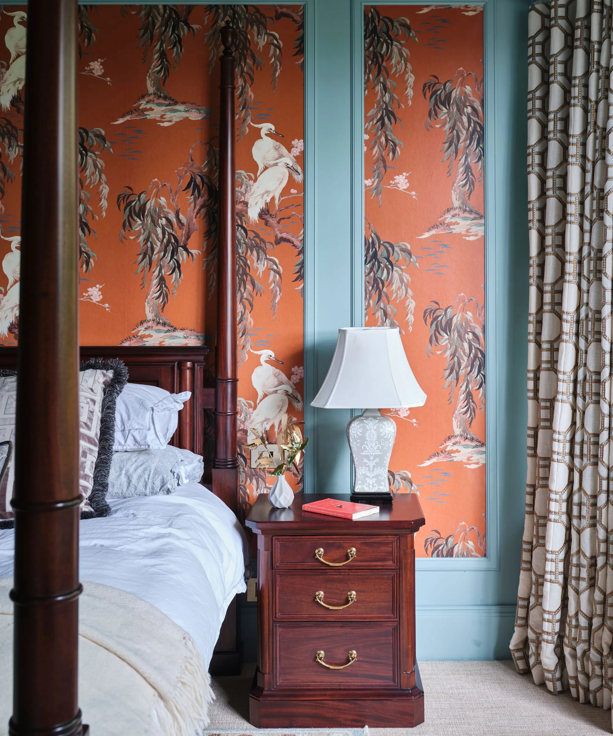 Ein Schlafzimmer mit blauen Wänden, getäfelter orangefarbener Statement-Tapete, einem Himmelbett aus dunklem Holz mit weißer Bettwäsche, einem passenden Nachttisch, weißen Vorhängen mit Lampenmuster und einem neutralen Teppich.' loading='lazy' title=