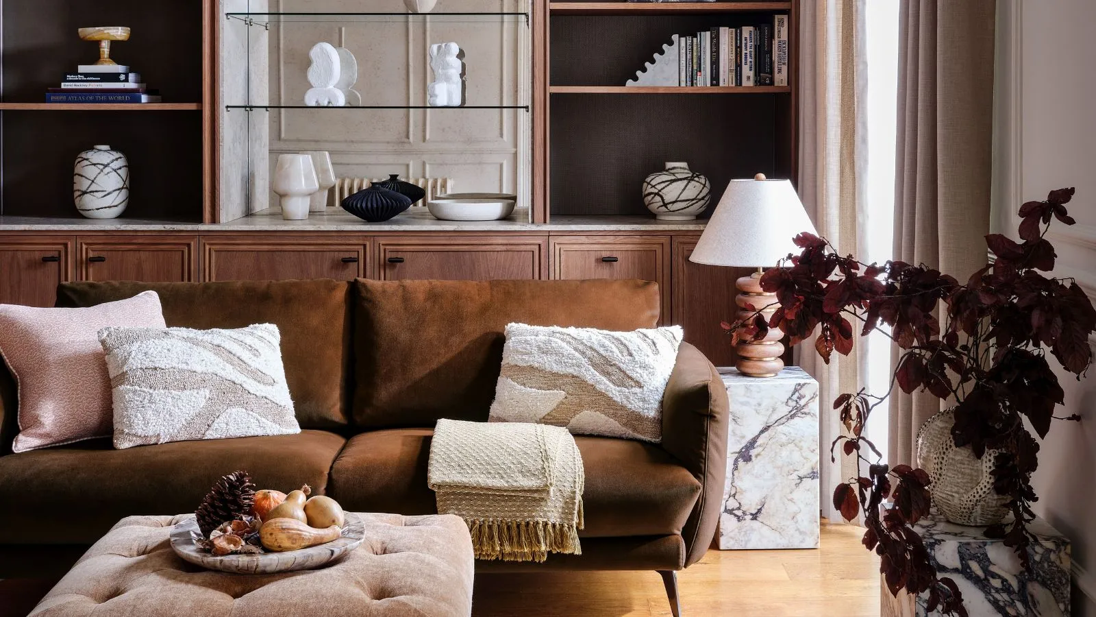 Um sofá de veludo marrom em uma sala neutra. Grandes prateleiras de madeira embutidas atrás de uma pequena mesa de mármore ao lado e uma planta de folhas marrons em primeiro plano.' loading='lazy' title=