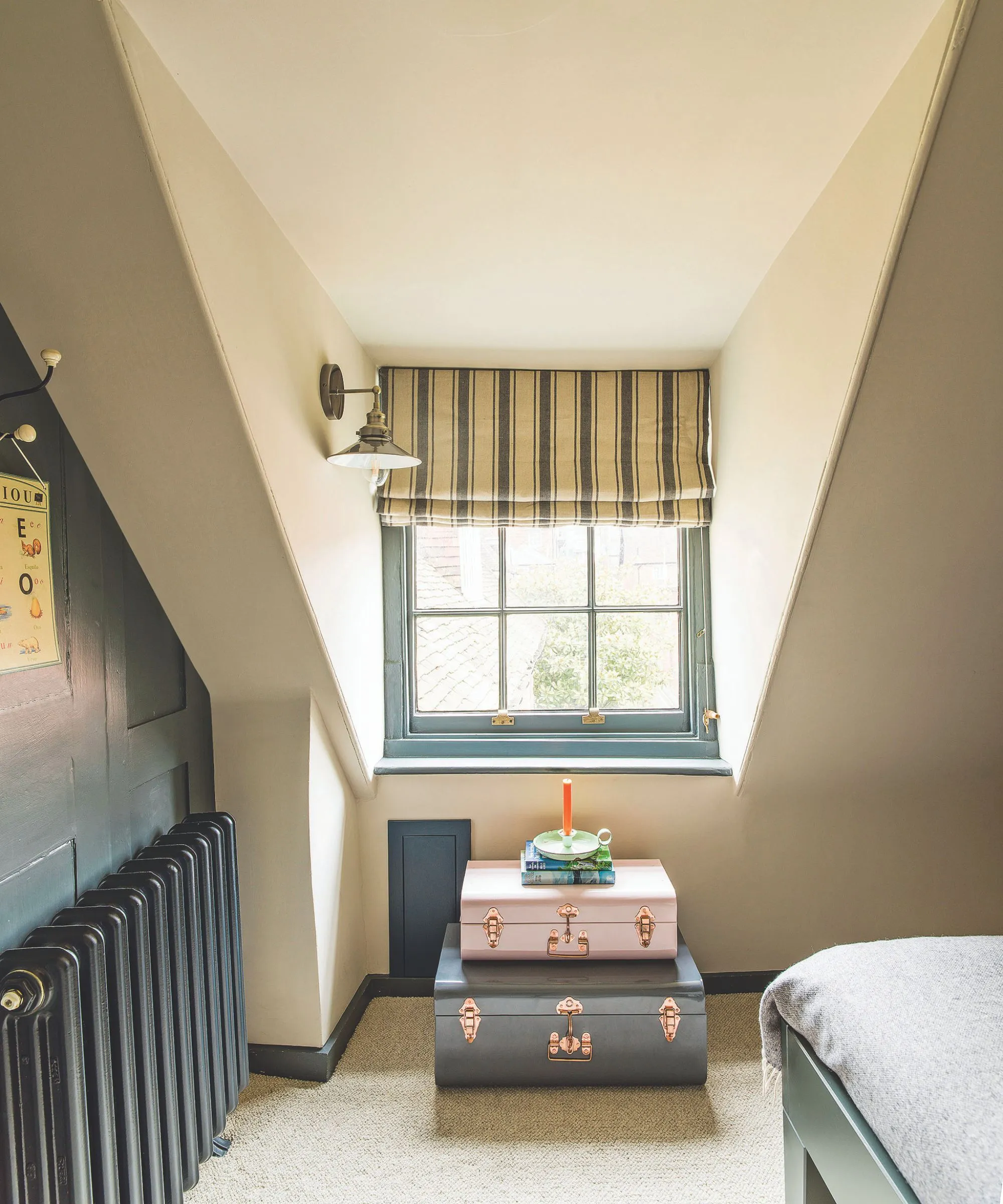 Una mansarda bianca con una parete verde acqua e un radiatore, una tenda a strisce bianche e blu e un set di valigie impilate. A destra c'è l'angolo di una struttura del letto blu con biancheria da letto grigia.' loading='lazy' title=