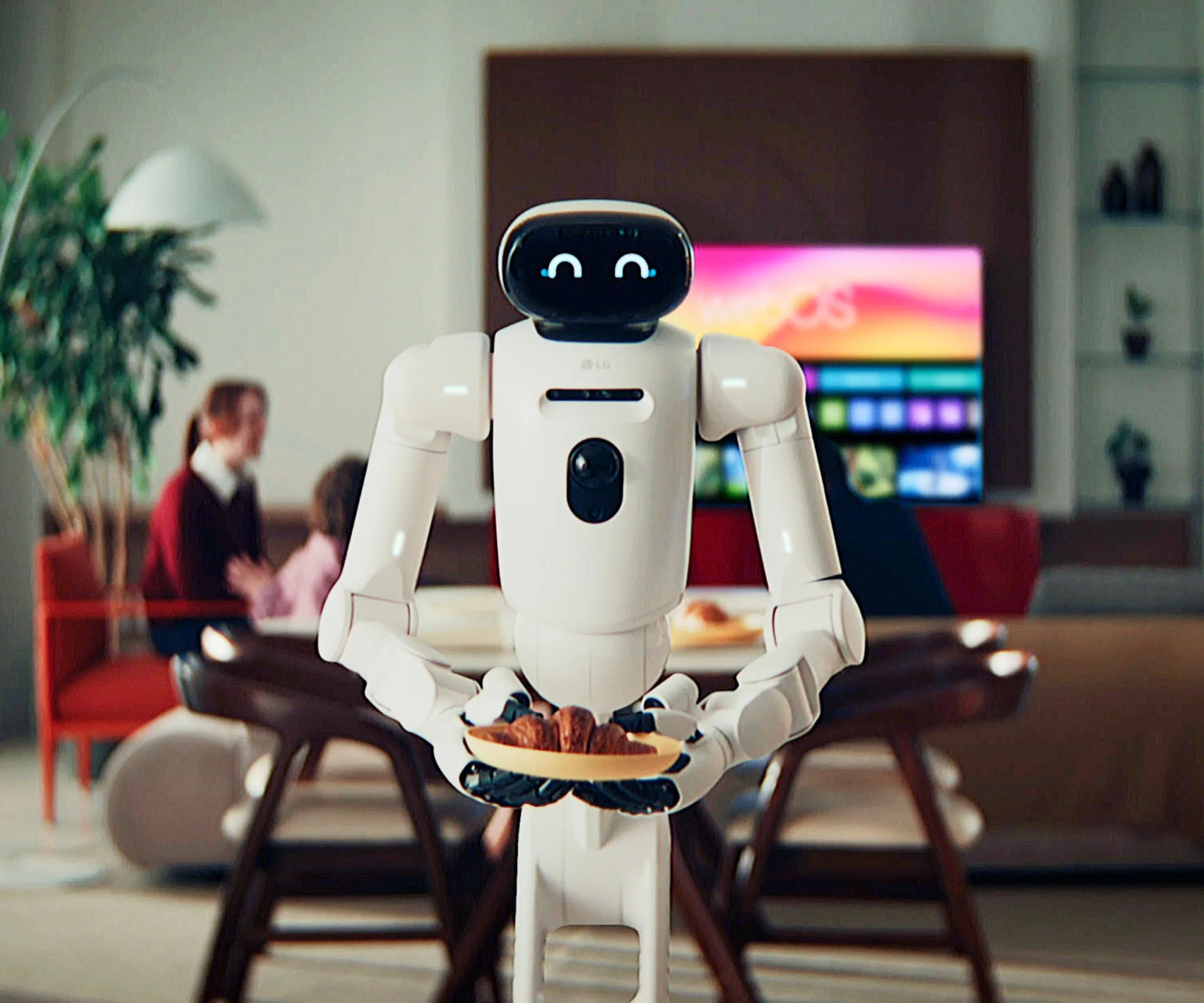 LG CLOiD trzymający rogalika na małym talerzu na zdjęciu przed rodziną siedział w salonie wokół telewizora.' loading='lazy' title=