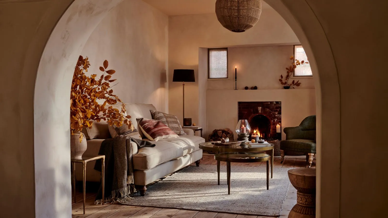 Przytulny salon w jesiennym stylu oglądany przez łukowe przejście. Wygodna kremowa sofa z zielonymi i czerwonymi poduszkami za okrągłym stolikiem kawowym. Obok w wazonie stoi duża kompozycja z pomarańczowych liści.' loading='lazy' title=
