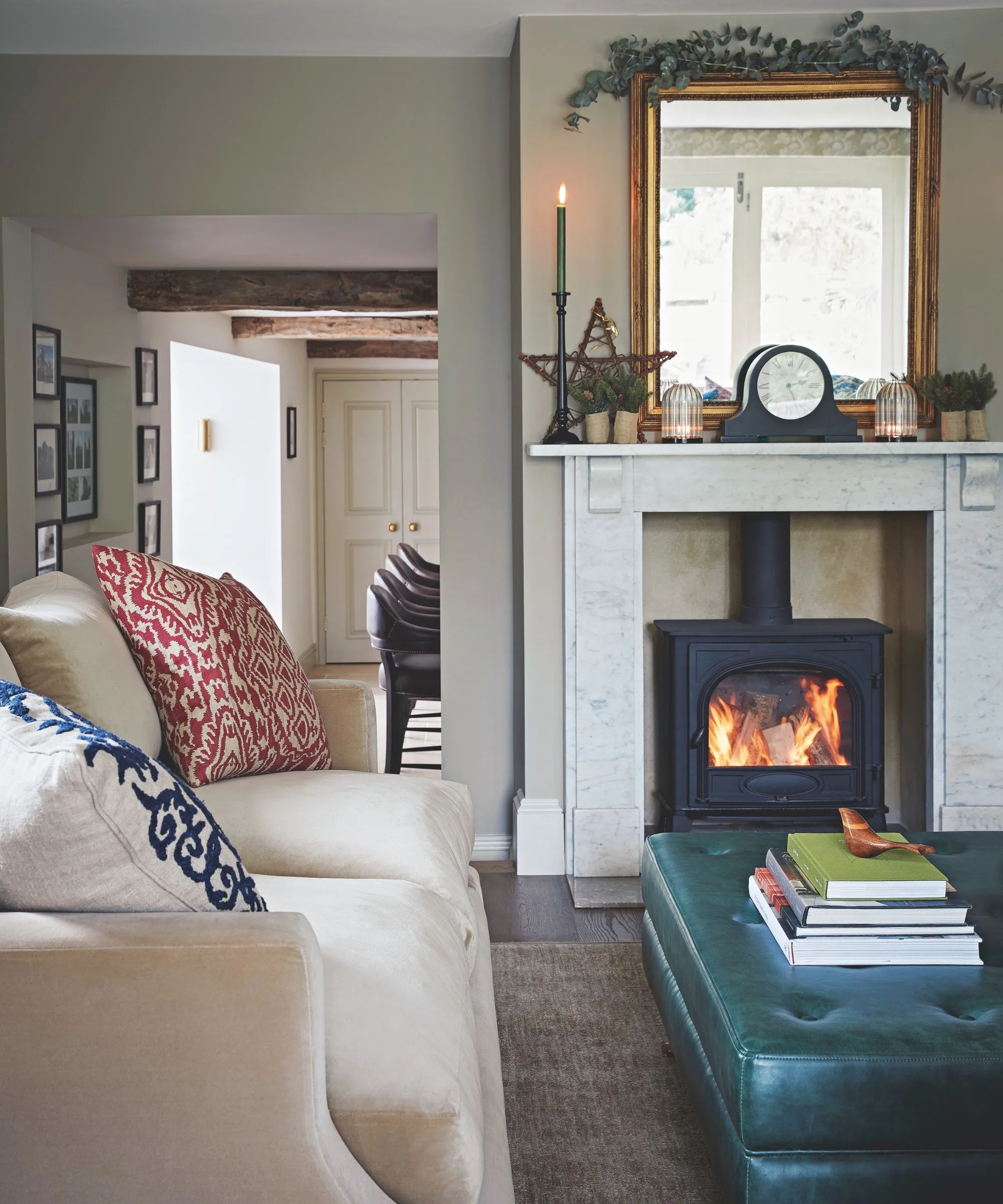 火のついた薪バーナー、テーパーキャンドル、白いソファ模様のクッション、革製のオットマン、大理石のマントルピース、その上に装飾的な時計と鏡が置かれた居心地の良いリビングルーム。' loading='lazy' title=