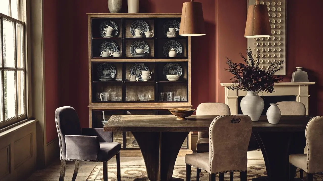 Uma sala de jantar em vermelho escuro com uma grande mesa de jantar de madeira. Cadeiras estofadas ao redor e dois pingentes de couro marrom pendurados acima. Um armário de madeira com louças finas ao fundo.' loading='lazy' title=