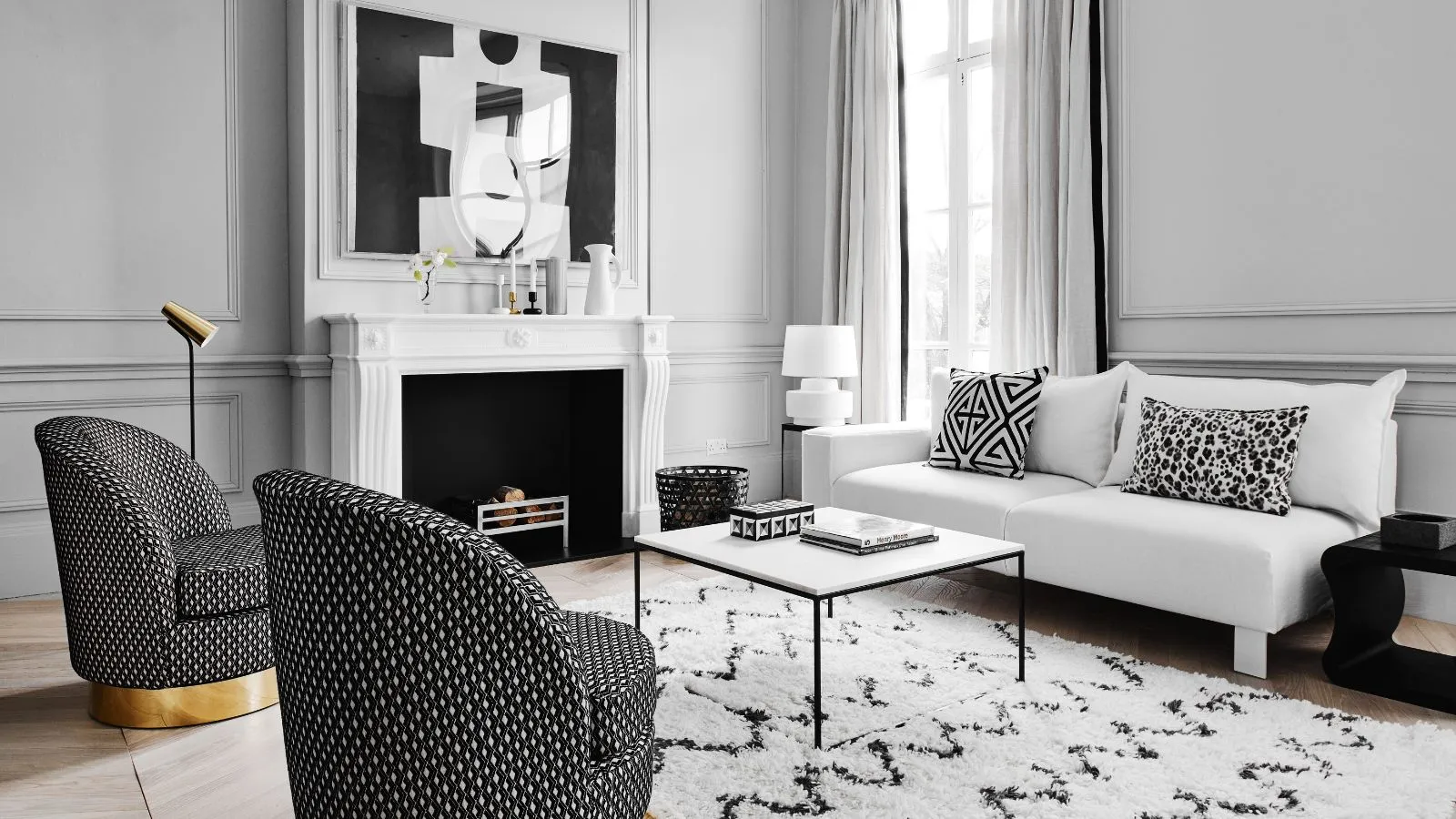 Una sala de estar en blanco y negro con paredes blancas, una repisa de chimenea blanca y cortinas blancas y una obra de arte en blanco y negro sobre la chimenea, una alfombra en blanco y negro, un sofá blanco con cojines en blanco y negro, una mesa de centro en blanco y negro y dos sillas en blanco y negro con bases doradas.' loading='lazy' title=