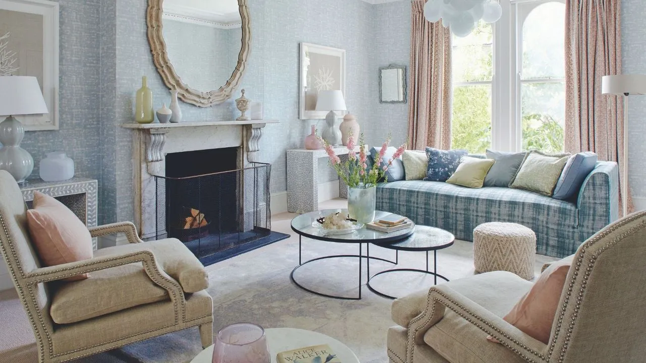 Una sala de estar en colores pastel con papel tapiz estampado en azul, una repisa de chimenea de mármol blanco con un espejo circular arriba y grandes ventanales a la derecha con cortinas de color rosa pastel. En primer plano hay dos sillas tapizadas en color crema y en el centro de la habitación hay una mesa de café circular con flores rosas y un sofá a cuadros azul detrás. En el suelo hay una alfombra de terciopelo blanco.' loading='lazy' title=