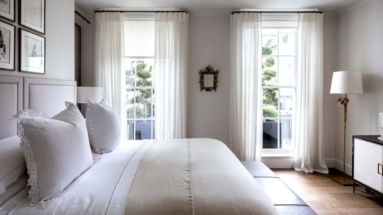 Un dormitorio blanco con una cama doble con ropa de cama blanca, paredes blancas, suelos de madera, una lámpara dorada con pantalla blanca, armarios blancos y negros y dos grandes ventanales con cortinas blancas transparentes.' loading='lazy' title=