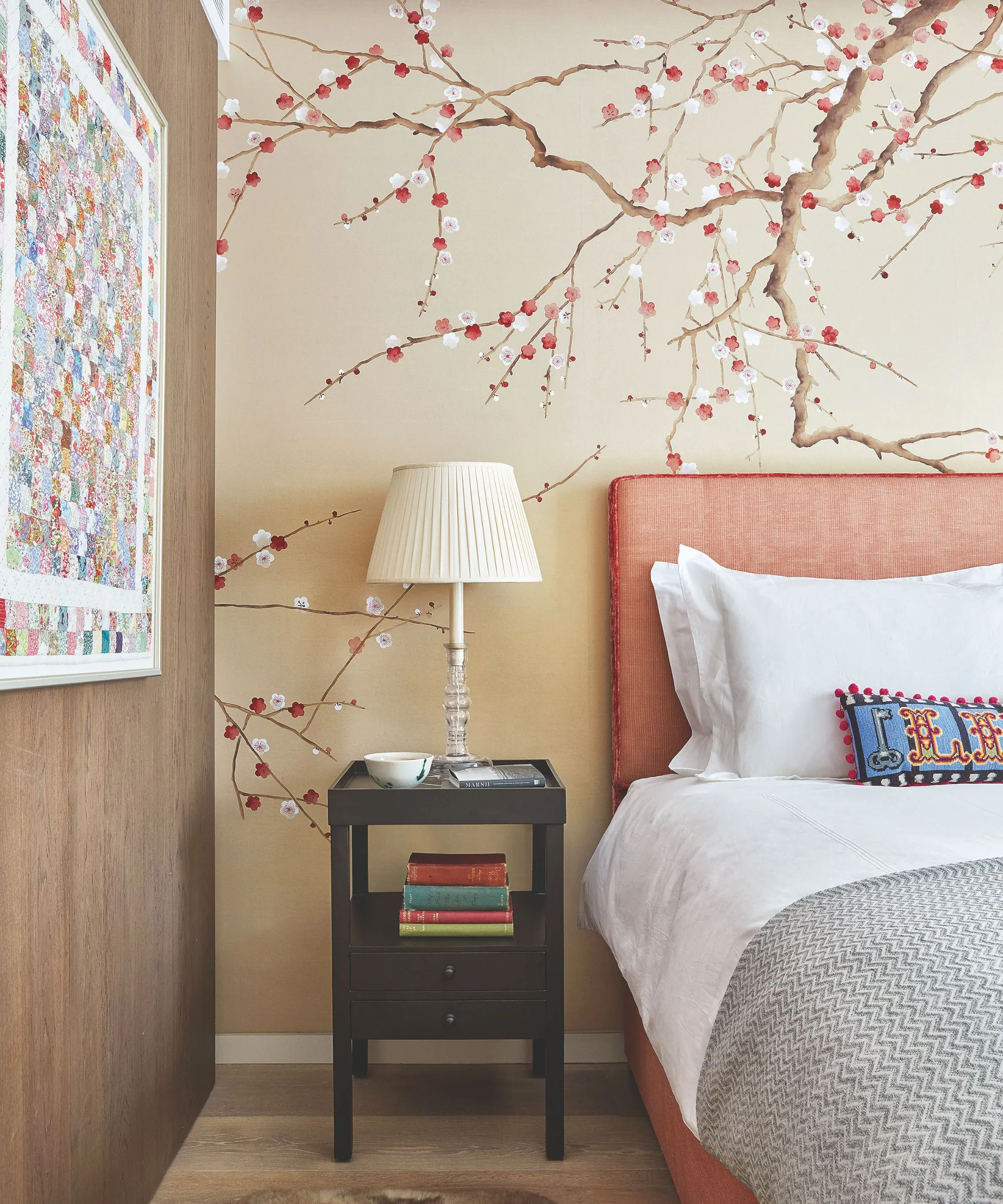 Un dormitorio con una cama naranja a la derecha con ropa de cama blanca, una manta gris y un cojín colorido. La pared detrás de la cama es de un color beige neutro con un motivo de cerezo en flor. A la izquierda de la cama hay una mesita de noche negra con una pila de libros y una lámpara blanca y a la izquierda hay una pared con paneles de madera con una obra de arte abstracta brillante y colorida. El suelo es de madera.' loading='lazy' title=