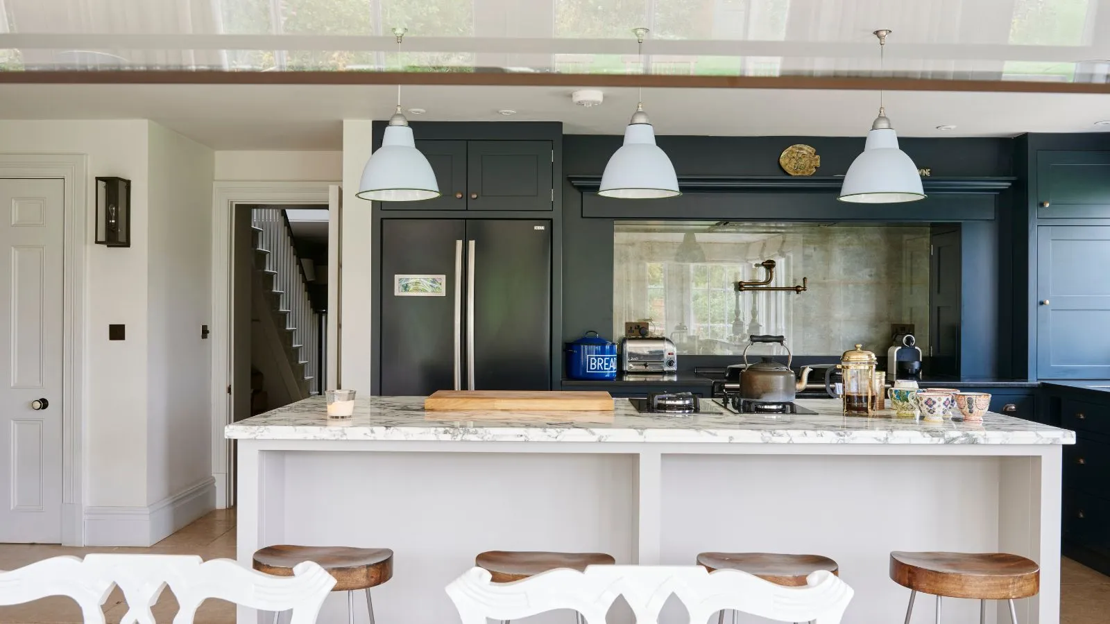 Una cucina luminosa e ariosa con un'isola di marmo bianca quattro sgabelli in legno scuro armadi e tre luci a sospensione bianca sospesa' loading='lazy' title=