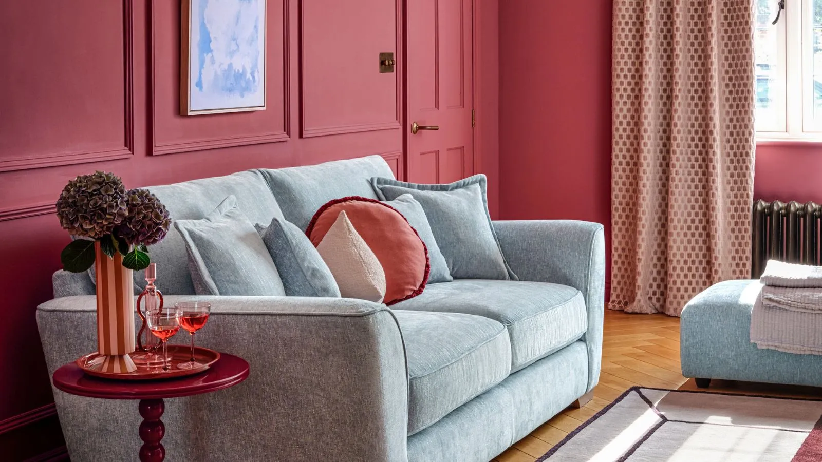 Un salon peint rouge avec un canapé bleu clair avec des coussins rouges dessus. Une petite table d'appoint rouge à côté du canapé avec un grand vase de fleurs dessus.' loading='lazy' title=