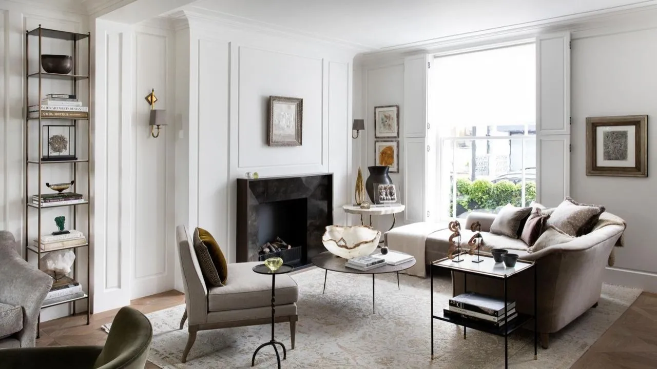 Un salon blanc avec des meubles neutres, de grandes fenêtres, des murs lambrissés blancs, un tapis à motifs de style persan pâle et une cheminée en marbre noir au centre du mur du fond. À gauche de la cheminée se trouve une grande étagère avec des livres et des décorations.' loading='lazy' title=
