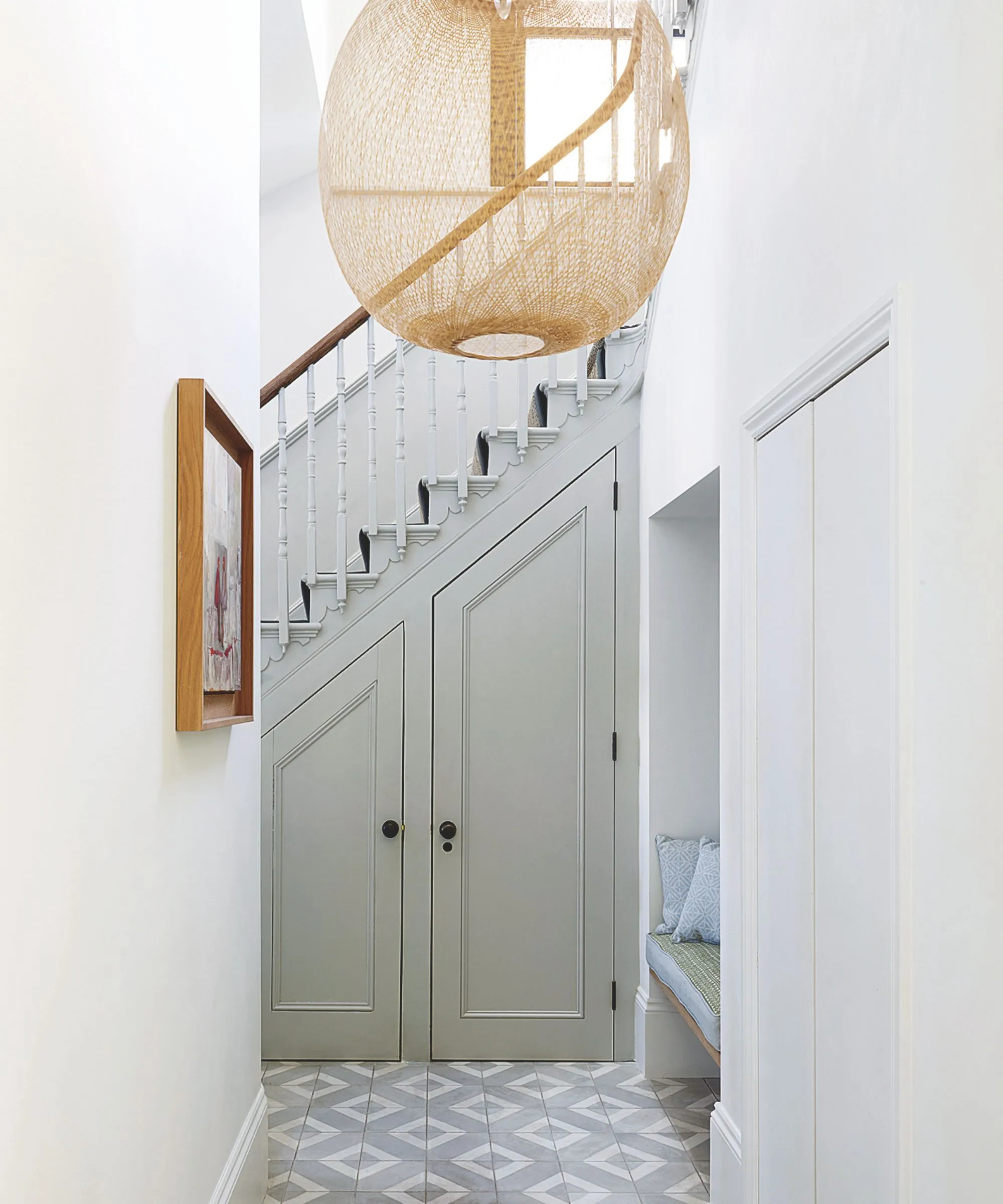 Une entrée blanche avec du carrelage au sol, une lampe en rotin, un banc intégré, une œuvre d'art encadrée en bois sur le mur et une grande lampe en rotin suspendue au-dessus.' loading='lazy' title=