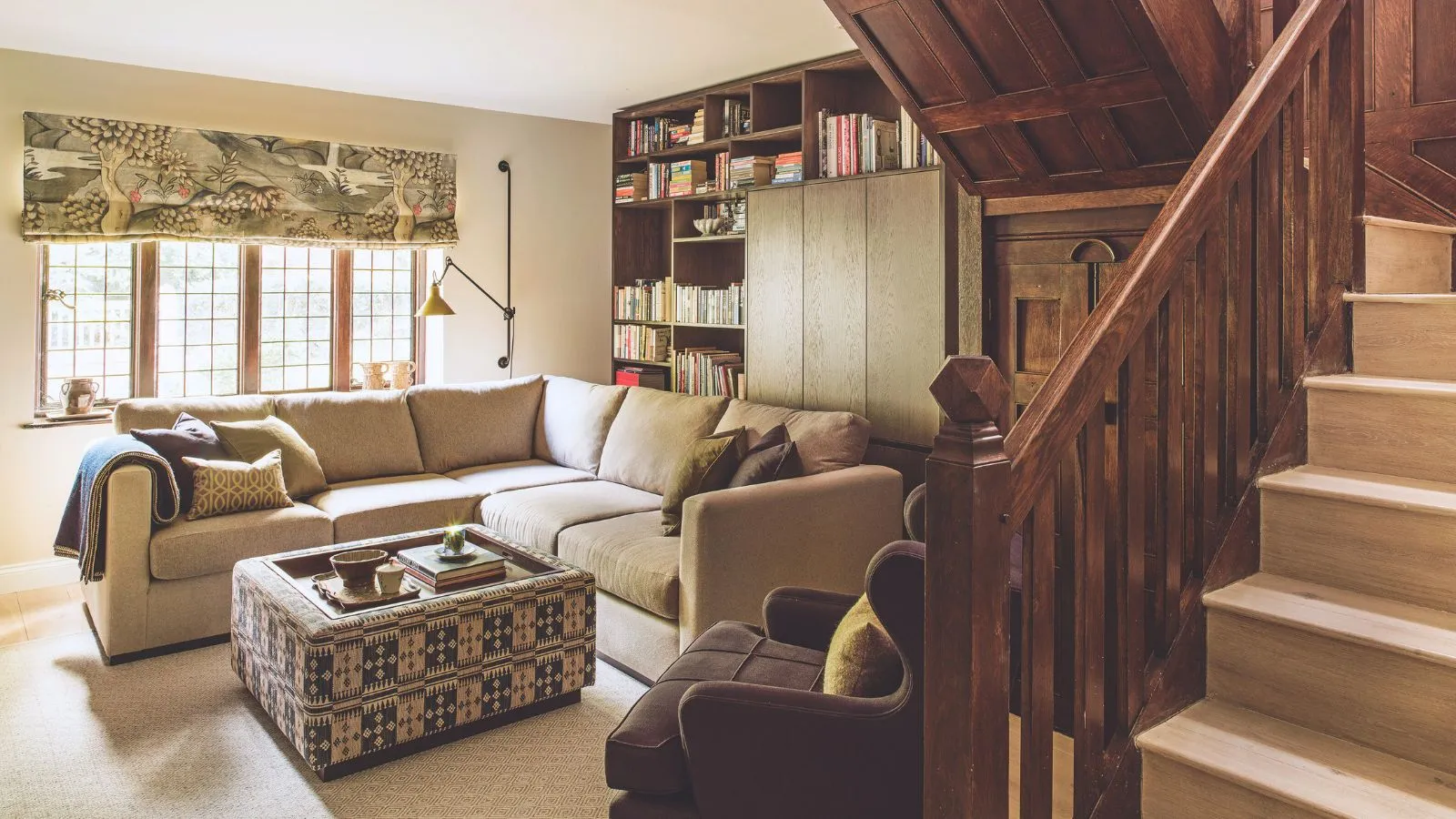 Et landingsområde med en trappe og træforbanister og et opholdsområde med en stor sofa og fodskammel. Monterede reoler.' loading='lazy' title=