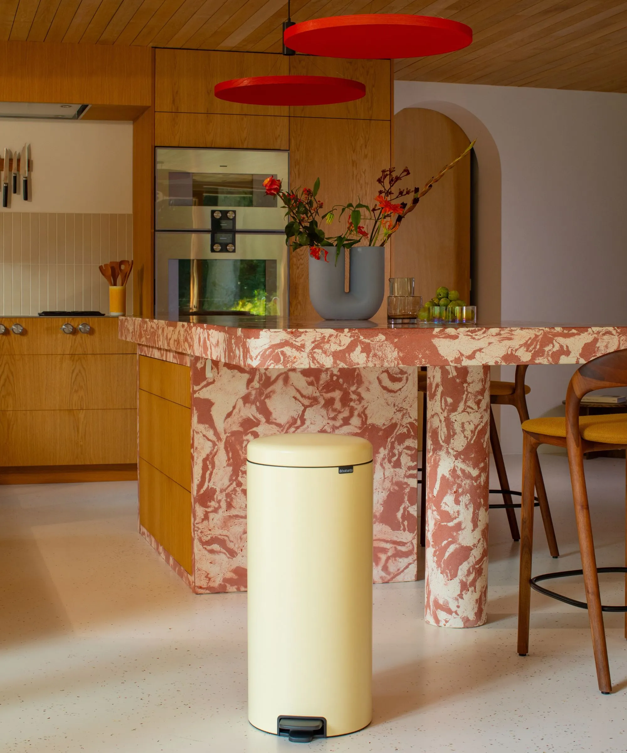 Una cocina de mármol y madera con un bote de basura de color amarillo mantequilla en primer plano, encimeras de mármol rojo y gabinetes de madera.' loading='lazy' title=