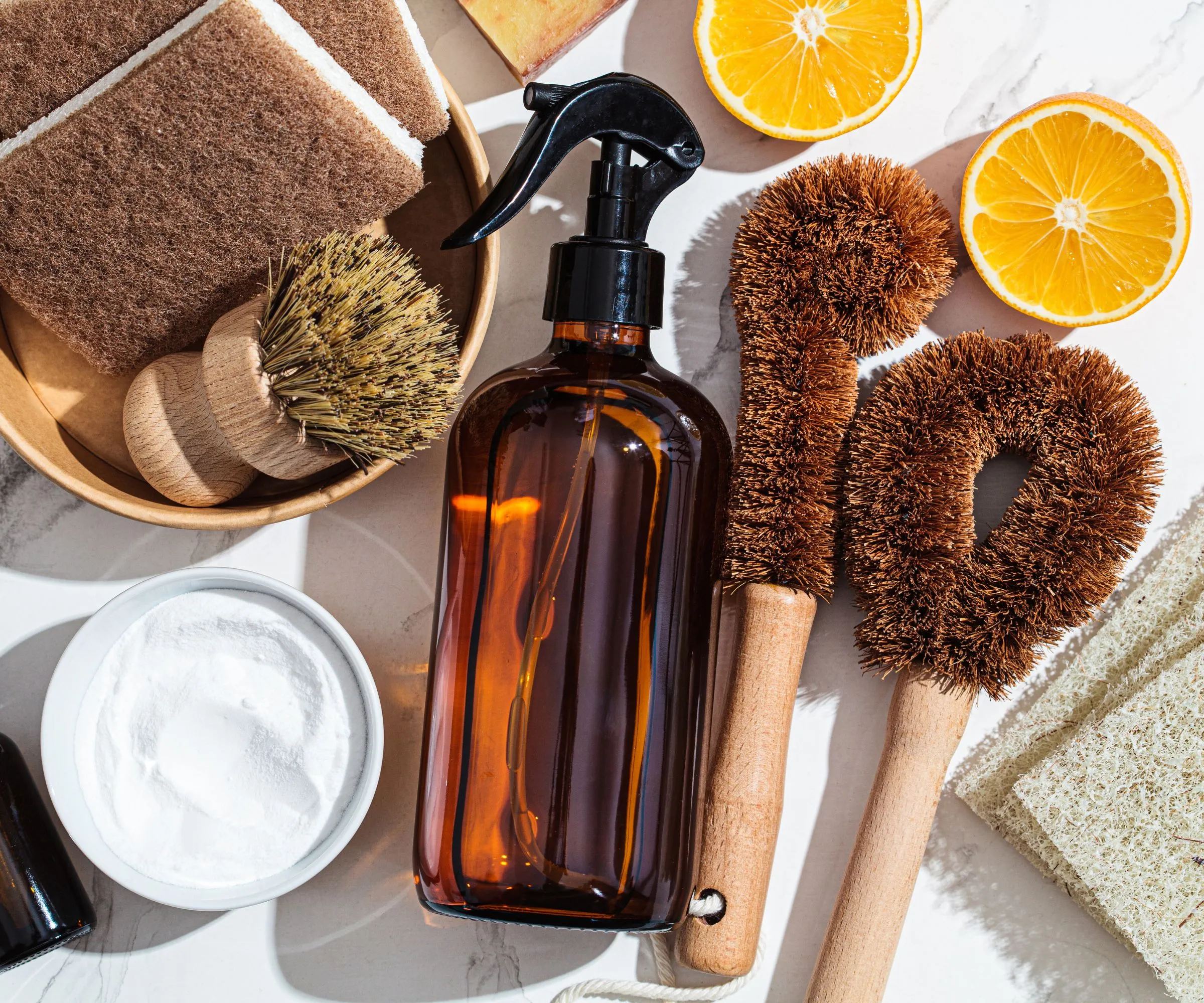 Una botella de vidrio ámbar rodeada de limones, bicarbonato de sodio y cepillos para fregar sobre una superficie de mármol.' loading='lazy' title=