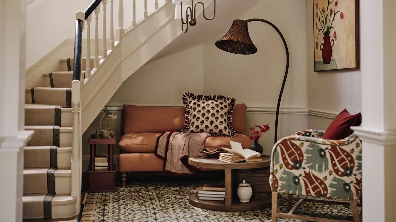 Un'accogliente area salotto con un piccolo divano e una poltrona annidata sotto una scala a chiocciola. Accanto a loro una grande lampada scultorea e una sospensione bassa.' loading='lazy' title=