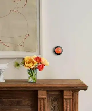 Ein kleiner orangefarbener Smart-Thermostat an einer beigen Wand über einer braunen, mit roten und gelben Blumen geschmückten Seite