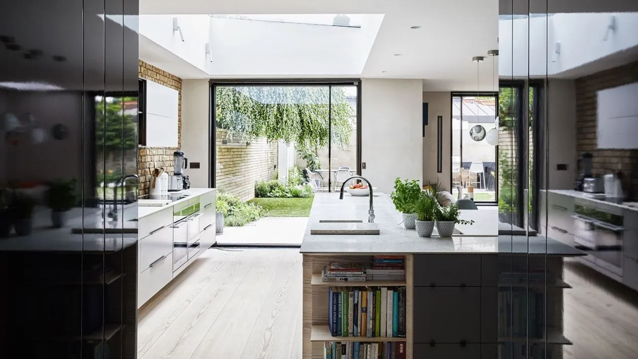 Une cuisine moderne, lumineuse et aérée avec du parquet, des armoires noires et blanches, un îlot avec des comptoirs en marbre blanc et des étagères intégrées remplies de livres de recettes, un dosseret en briques apparentes et de grandes portes-fenêtres donnant sur un jardin verdoyant.' loading='lazy' title=