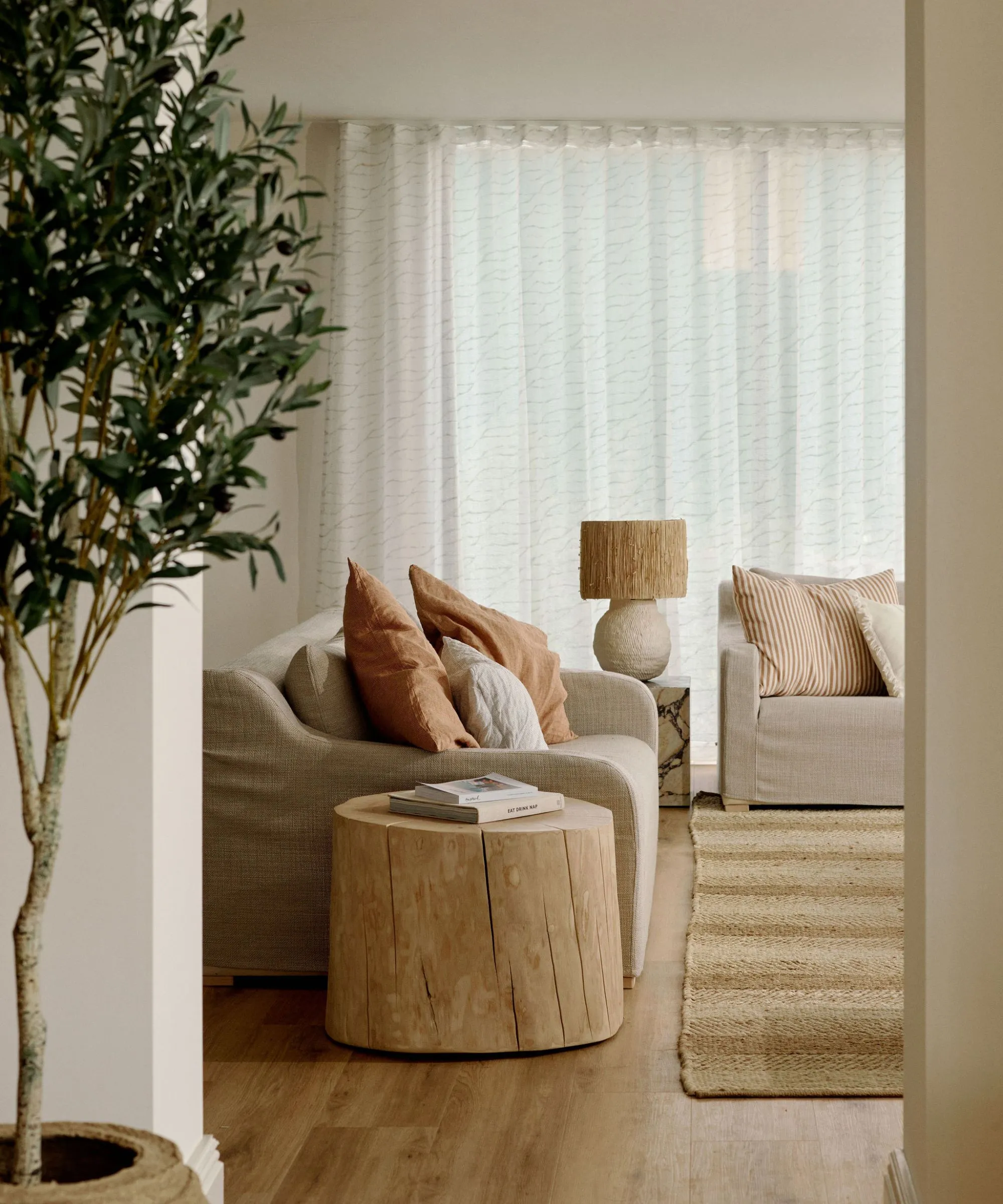 neutral varm minimalistisk stue med et jute tæppe to linned glidebelagte sofaer et rustikt træ sidebord og et oliventræ' loading='lazy' title=