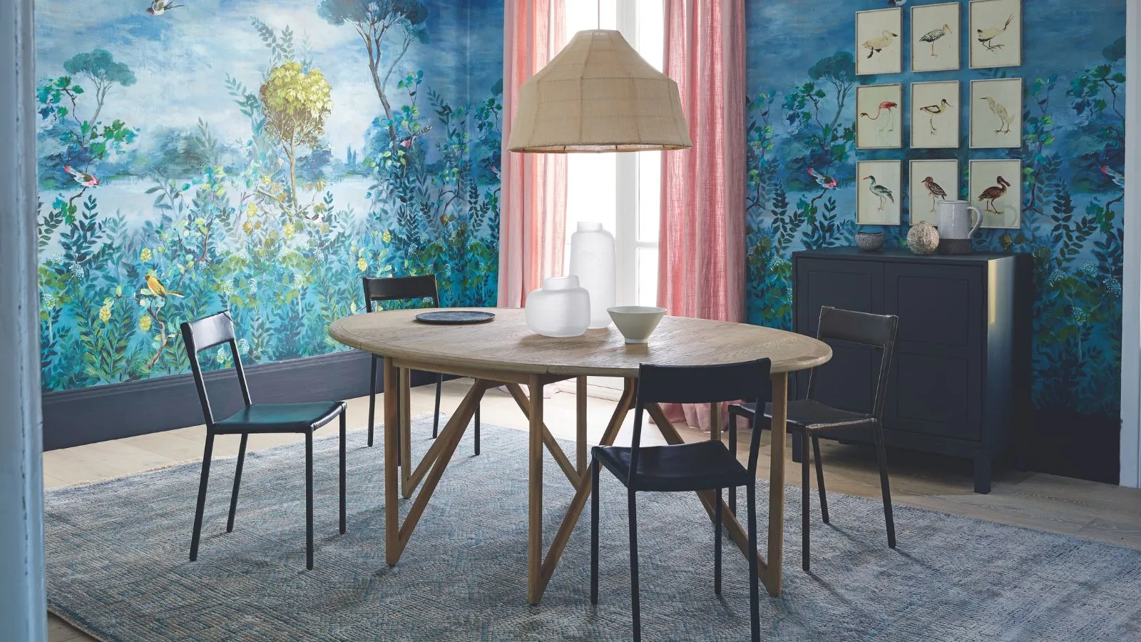 Uma sala de jantar azul com papel de parede estampado, cortinas rosa, abajur de vime, mesa redonda de madeira e cadeiras pretas.' loading='lazy' title=