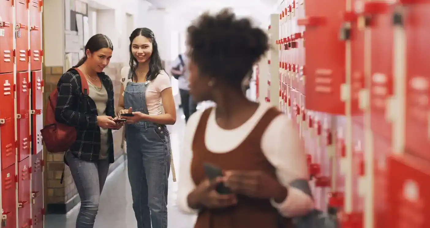 Teen girl victim of social bullying looks back on two bullies in the school hallway. All three girls hold cell phones.